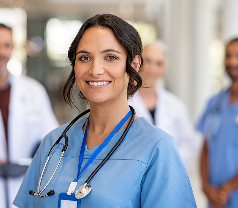 Portrait of happy young nurse in uniform with healthcare team in background. Successful team of doctor and nurses smiling. Beautiful and satisfied healthcare worker in private clinic looking at camera